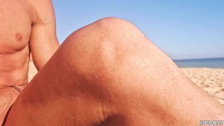 Sexy guy shows off his hard cock on a public beach with people walking by - girl gives him a handjob